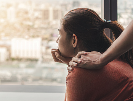 Woman staring out window leaning on her chin, with someone's arm on her shoulder